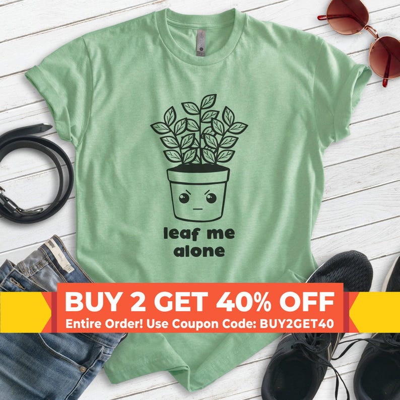 Op de afbeelding: Een groen T-shirt met een zwart-witte illustratie van een plant in een pot met een gezicht. De plant heeft een chagrijnige uitdrukking en de tekst "leaf me alone" staat onder de plant gedrukt. Het T-shirt heeft een aanbieding van twee voor 40% korting.