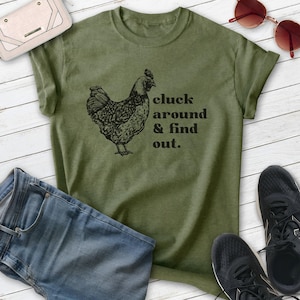 Może przedstawiać: Oliwkowo-zielona koszulka z czarną ilustracją kury i napisem "cluck around & find out". Na zdjęciu znajdują się niebieskie dżinsy, czarne trampki, kremowy portfel i czerwone okulary przeciwsłoneczne.