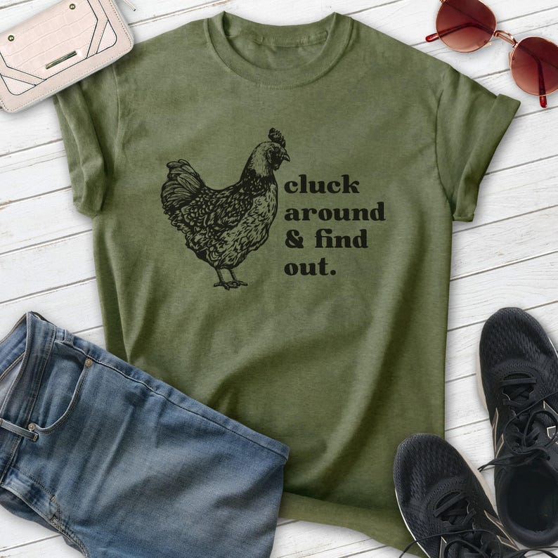 Może przedstawiać: Oliwkowo-zielona koszulka z czarną ilustracją kury i napisem "cluck around & find out". Na zdjęciu znajdują się niebieskie dżinsy, czarne trampki, kremowy portfel i czerwone okulary przeciwsłoneczne.