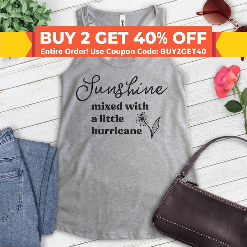 Puede incluir: Camiseta de tirantes gris con el texto "Sunshine mixed with a little hurricane" en letras negras. Un diente de le&oacute;n con tallo y hojas est&aacute; al lado del texto.