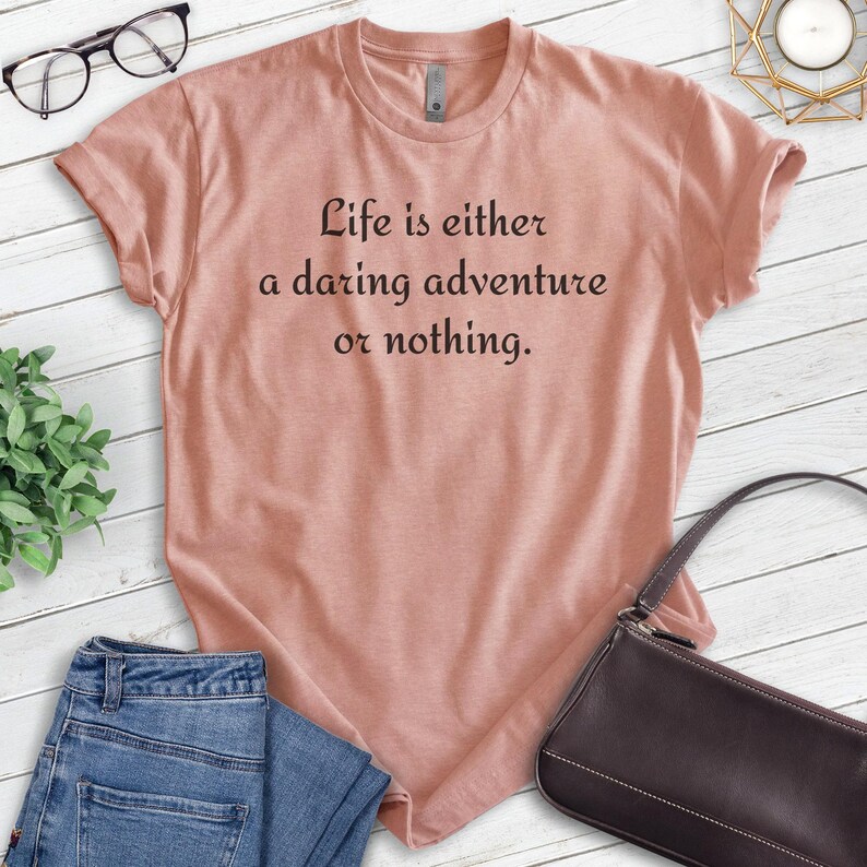 May include: A light pink t-shirt with the text "Life is either a daring adventure or nothing." printed in black.