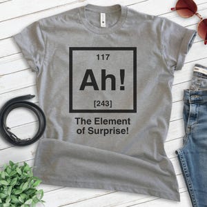 Pode incluir: Camiseta cinza com um gr&aacute;fico quadrado preto que diz "Ah!" com o n&uacute;mero 117 acima e [243] abaixo. O texto abaixo do quadrado diz "The Element of Surprise!"