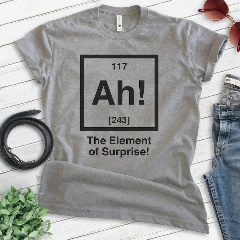 Pode incluir: Camiseta cinza com um gr&aacute;fico quadrado preto que diz "Ah!" com o n&uacute;mero 117 acima e [243] abaixo. O texto abaixo do quadrado diz "The Element of Surprise!"