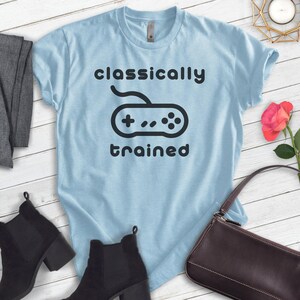 May include: Light blue t-shirt with a black graphic of a video game controller and the text "classically trained".