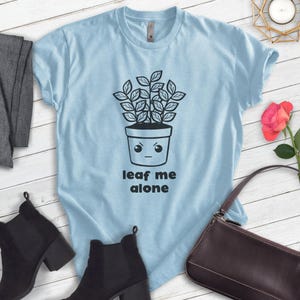 Op de afbeelding: Lichtblauw T-shirt met een zwart-witte illustratie van een plant in een pot met een gezicht. De plant heeft een chagrijnige uitdrukking en de tekst "leaf me alone" is onder de plant gedrukt.