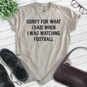 Pode incluir: Uma camiseta cinza claro com o texto "Desculpe pelo que eu disse quando estava assistindo futebol" impresso em preto.