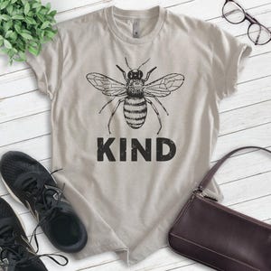 May include: A light gray t-shirt with a black and white illustration of a bee and the word "KIND" printed on the front.