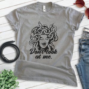Pu&ograve; includere: T-shirt grigio con un'illustrazione nera di Medusa e la scritta "Don't look at me". La maglietta &egrave; abbinata a jeans blu, occhiali da sole e una cintura nera. La maglietta &egrave; realizzata in materiale morbido.