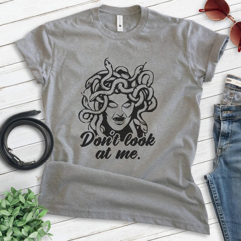 Pu&ograve; includere: T-shirt grigio con un'illustrazione nera di Medusa e la scritta "Don't look at me". La maglietta &egrave; abbinata a jeans blu, occhiali da sole e una cintura nera. La maglietta &egrave; realizzata in materiale morbido.