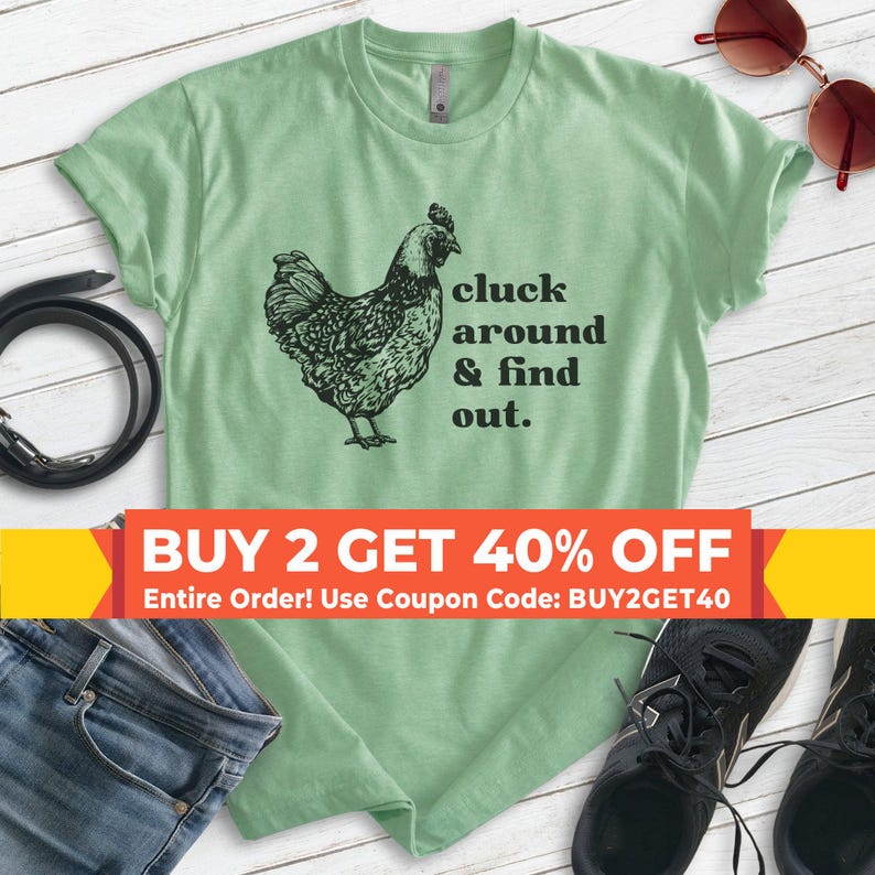 Może przedstawiać: Jasnozielona koszulka z czarną ilustracją kurczaka i napisem "cluck around & find out." Na zdjęciu widoczne są r&oacute;wnież okulary przeciwsłoneczne, pasek, dżinsy i trampki.
