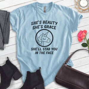Puede incluir: Camiseta azul claro con el texto "She's Beauty She's Grace She'll Stab You In The Face" y un gr&aacute;fico de unicornio en blanco y negro. La camiseta se muestra con botines negros, un su&eacute;ter gris y un bolso marr&oacute;n.