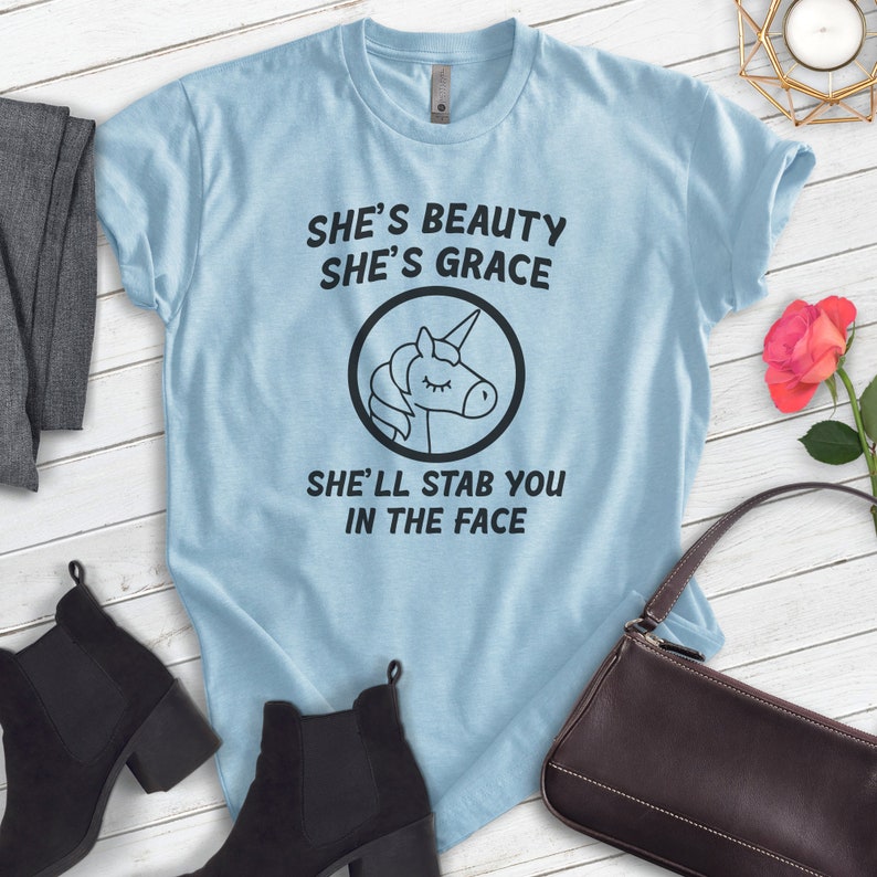 Puede incluir: Camiseta azul claro con el texto "She's Beauty She's Grace She'll Stab You In The Face" y un gr&aacute;fico de unicornio en blanco y negro. La camiseta se muestra con botines negros, un su&eacute;ter gris y un bolso marr&oacute;n.
