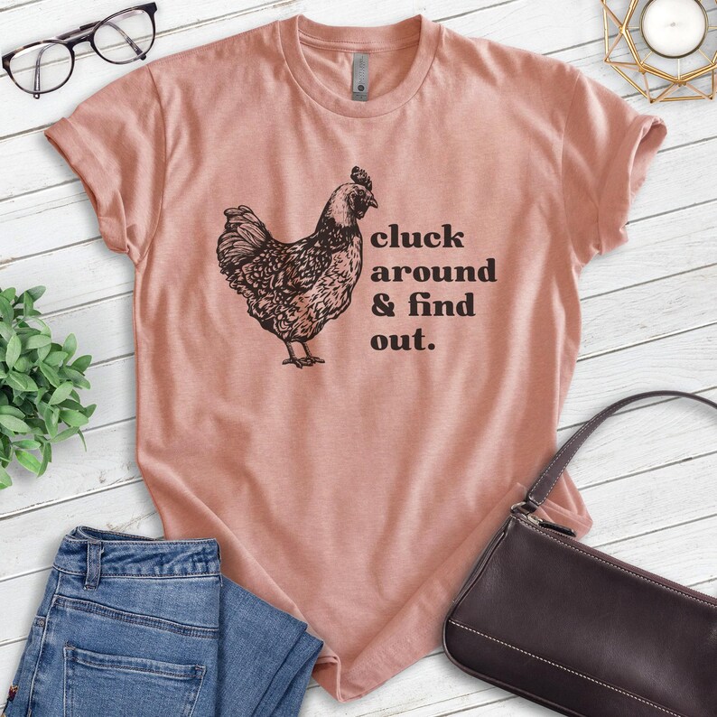 Może przedstawiać: Jasnor&oacute;żowa koszulka z czarną ilustracją kury i napisem "cluck around & find out." Koszulka jest zestawiona z niebieskimi dżinsami i czarną torebką.