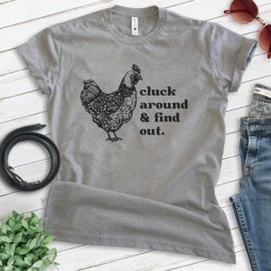 Może przedstawiać: Szara koszulka z czarną ilustracją kury i napisem "cluck around & find out". Ta koszulka to casualowy element garderoby, odpowiedni do noszenia na co dzień. Projekt jest wydrukowany w kolorze czarnym.