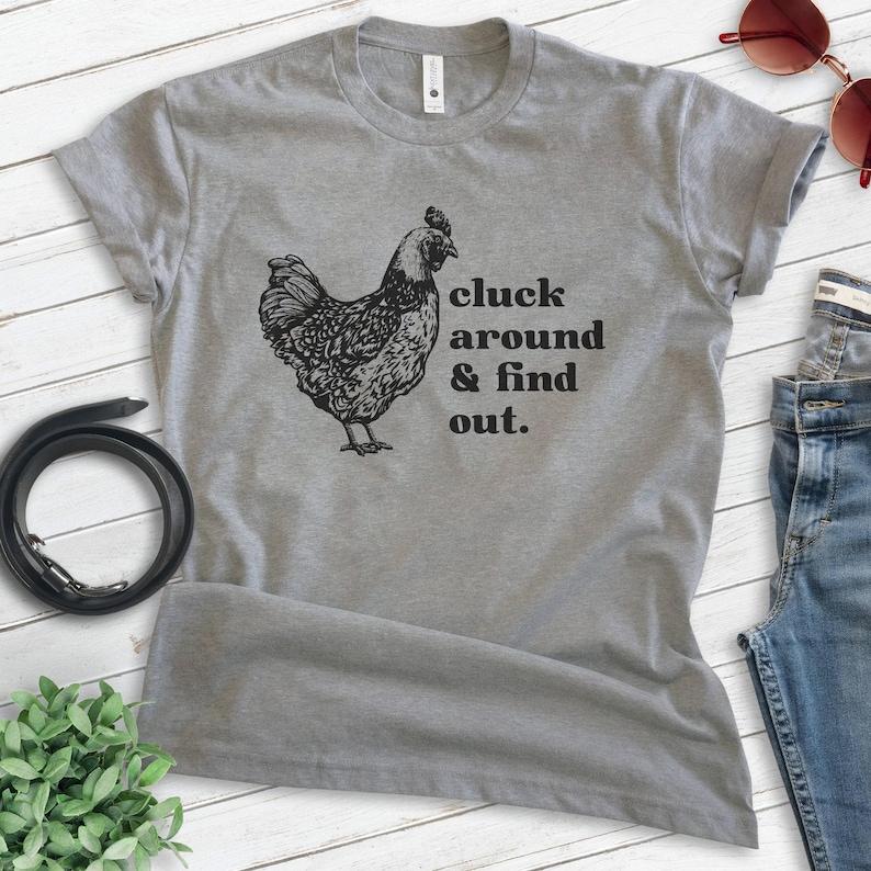 Może przedstawiać: Szara koszulka z czarną ilustracją kury i napisem "cluck around & find out". Ta koszulka to casualowy element garderoby, odpowiedni do noszenia na co dzień. Projekt jest wydrukowany w kolorze czarnym.