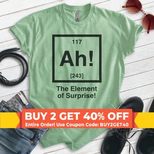 Pode incluir: Uma camiseta verde com um gr&aacute;fico em preto e branco de um elemento da tabela peri&oacute;dica. O elemento &eacute; rotulado como "Ah!" com o n&uacute;mero at&ocirc;mico 117 e o peso at&ocirc;mico 243. O texto abaixo do elemento diz "The Element of Surprise!" A camiseta tem um fundo branco e o texto est&aacute; em preto. A imagem tamb&eacute;m inclui uma faixa amarela com o texto "BUY 2 GET 40% OFF" e "Entire Order! Use Coupon Code: BUY2GET40".