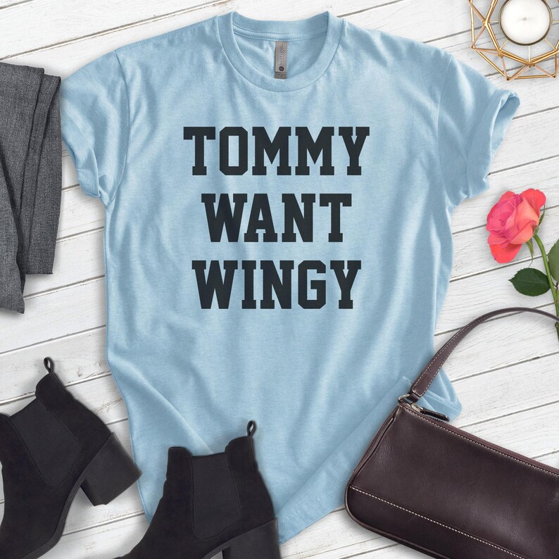 May include: Light blue t-shirt with the text "TOMMY WANT WINGY" in bold, black letters. The shirt is displayed on a white wooden surface, with black ankle boots, a gray garment, and a brown purse nearby.