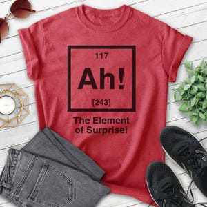 Pode incluir: Camiseta vermelha com um gr&aacute;fico quadrado preto que diz "Ah!" com o n&uacute;mero 117 acima e [243] abaixo. O texto abaixo do quadrado diz "The Element of Surprise!"
