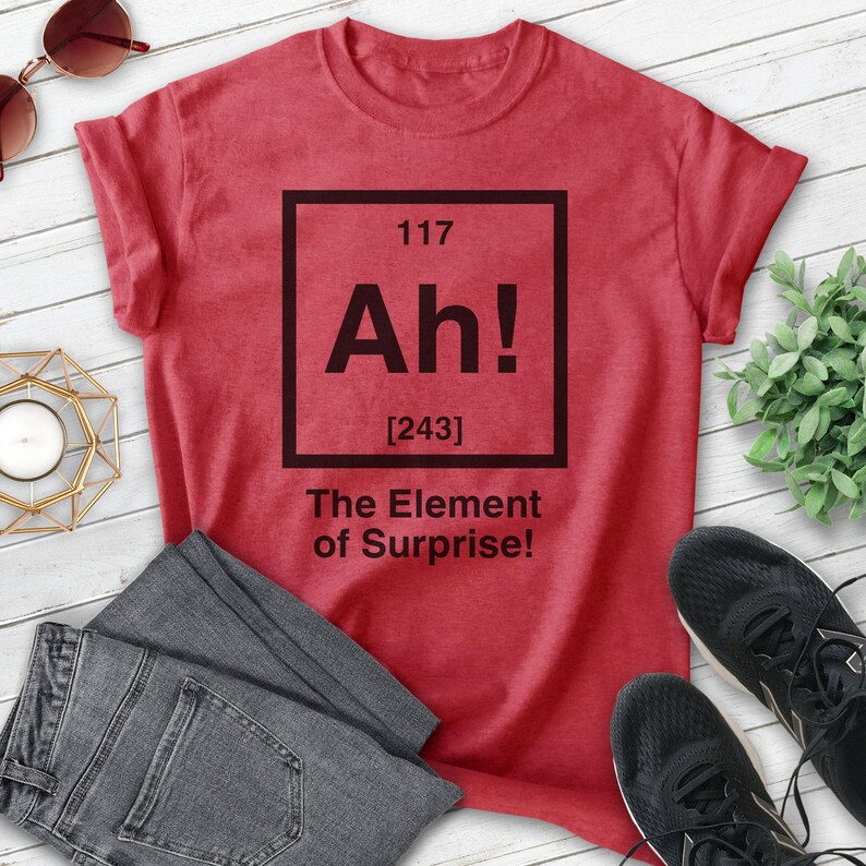 Pode incluir: Camiseta vermelha com um gr&aacute;fico quadrado preto que diz "Ah!" com o n&uacute;mero 117 acima e [243] abaixo. O texto abaixo do quadrado diz "The Element of Surprise!"