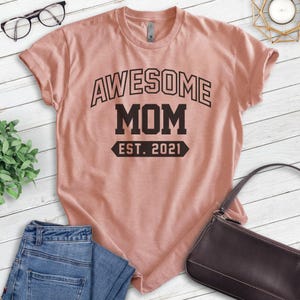 Peut inclure: Un t-shirt rose p&acirc;le avec le texte "Awesome Mom Est. 2021" en noir.