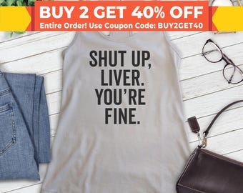Puede incluir: Un top de tirantes gris con el texto "SHUT UP, LIVER. YOU'RE FINE." impreso en negro. El top está sobre una superficie de madera blanca.