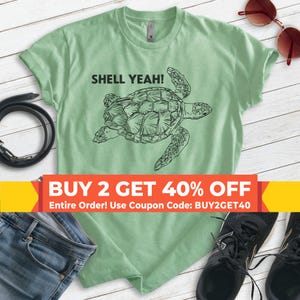 May include: A light green t-shirt with the text "SHELL YEAH!" and a black illustration of a sea turtle. The shirt is displayed with blue jeans, sunglasses, and black shoes.