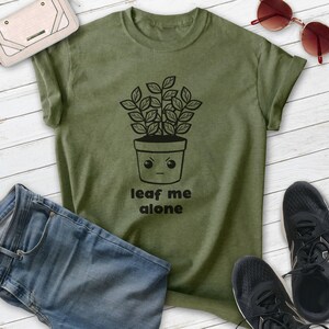 Op de afbeelding: Een groen T-shirt met een zwarte afbeelding van een plant in een pot met een gezicht waarop "leaf me alone" staat.