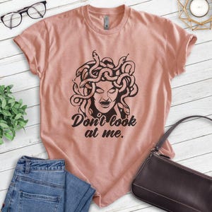 Pu&ograve; includere: T-shirt rosa chiaro con grafica nera di Medusa con serpenti al posto dei capelli e la scritta "Don't look at me." La maglietta ha maniche corte e girocollo.