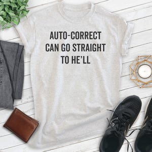 May include: A light gray t-shirt with the text "AUTO-CORRECT CAN GO STRAIGHT TO HE'LL" in black. The shirt is displayed with a brown wallet, black sneakers, and a pair of gray pants.