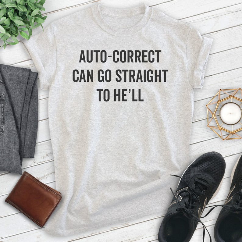 May include: A light gray t-shirt with the text "AUTO-CORRECT CAN GO STRAIGHT TO HE'LL" in black. The shirt is displayed with a brown wallet, black sneakers, and a pair of gray pants.