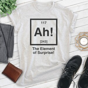 Pode incluir: Uma camiseta cinza com um gr&aacute;fico quadrado preto que diz "Ah! [243] The Element of Surprise!" O gr&aacute;fico &eacute; estilizado como um elemento da tabela peri&oacute;dica.