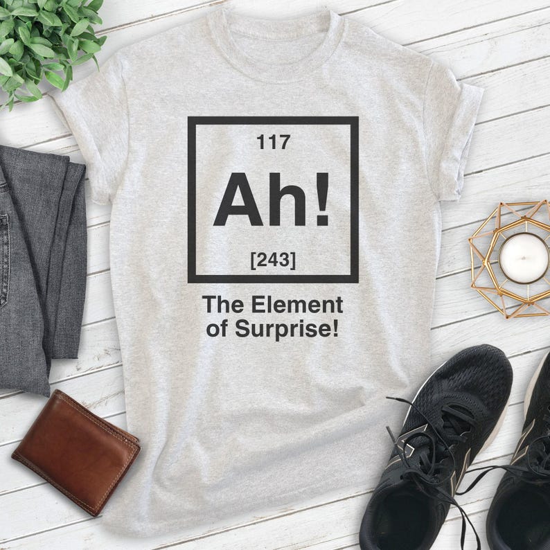 Pode incluir: Uma camiseta cinza com um gr&aacute;fico quadrado preto que diz "Ah! [243] The Element of Surprise!" O gr&aacute;fico &eacute; estilizado como um elemento da tabela peri&oacute;dica.