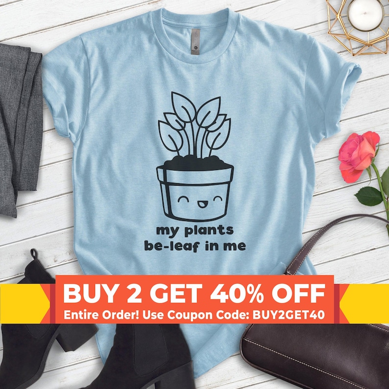 K&ouml;nnte beinhalten: Hellblaues T-Shirt mit einer schwarz-wei&szlig;en Illustration einer Topfpflanze mit einem l&auml;chelnden Gesicht. Der Text "my plants be-leaf in me" ist unter der Illustration gedruckt. Der Text "BUY 2 GET 40% OFF Entire Order! Use Coupon Code: BUY2GET40" ist unten im Bild gedruckt.