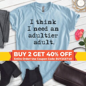 May include: Light blue t-shirt with the text "I think I need an adultier adult." in black typewriter font. The shirt is on a wooden surface with a pair of black boots, a black purse, and a rose.
