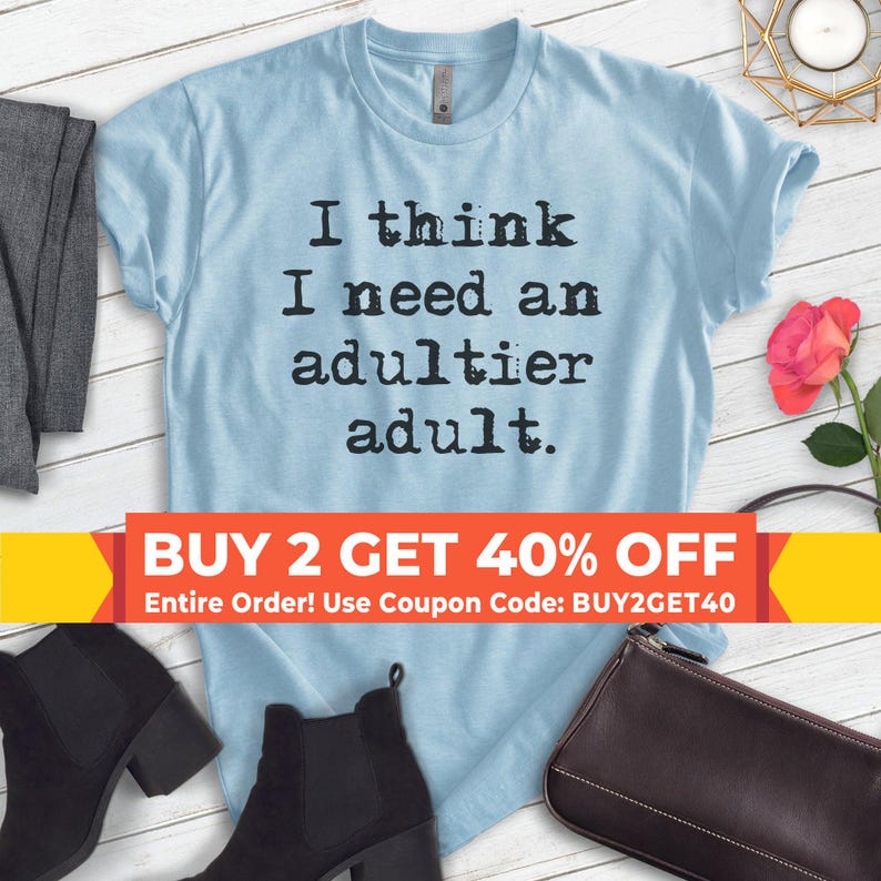 May include: Light blue t-shirt with the text "I think I need an adultier adult." in black typewriter font. The shirt is on a wooden surface with a pair of black boots, a black purse, and a rose.
