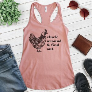 Pu&ograve; includere: Un top rosa con un'illustrazione nera di una gallina e il testo "cluck around & find out."