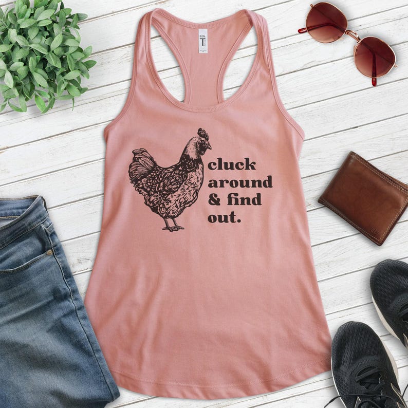 Pu&ograve; includere: Un top rosa con un'illustrazione nera di una gallina e il testo "cluck around & find out."