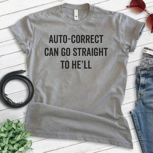 May include: A heather gray t-shirt with the text "AUTO-CORRECT CAN GO STRAIGHT TO HE'LL" in black. The shirt is displayed on a light wood-grain surface, with a black belt, sunglasses, and jeans visible.