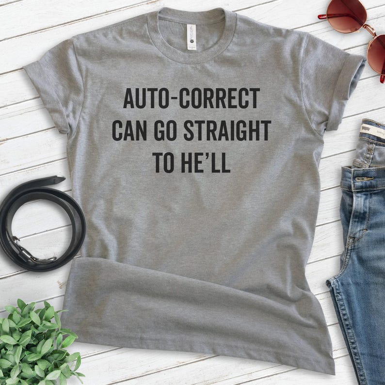 May include: A heather gray t-shirt with the text "AUTO-CORRECT CAN GO STRAIGHT TO HE'LL" in black. The shirt is displayed on a light wood-grain surface, with a black belt, sunglasses, and jeans visible.