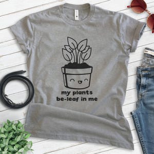 K&ouml;nnte beinhalten: Graues T-Shirt mit einer schwarz-wei&szlig;en Illustration einer Topfpflanze mit einem l&auml;chelnden Gesicht. Der Text "my plants be-leaf in me" ist unter der Illustration gedruckt.