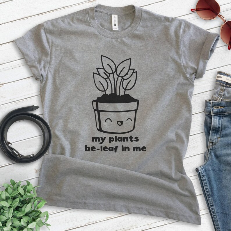 K&ouml;nnte beinhalten: Graues T-Shirt mit einer schwarz-wei&szlig;en Illustration einer Topfpflanze mit einem l&auml;chelnden Gesicht. Der Text "my plants be-leaf in me" ist unter der Illustration gedruckt.