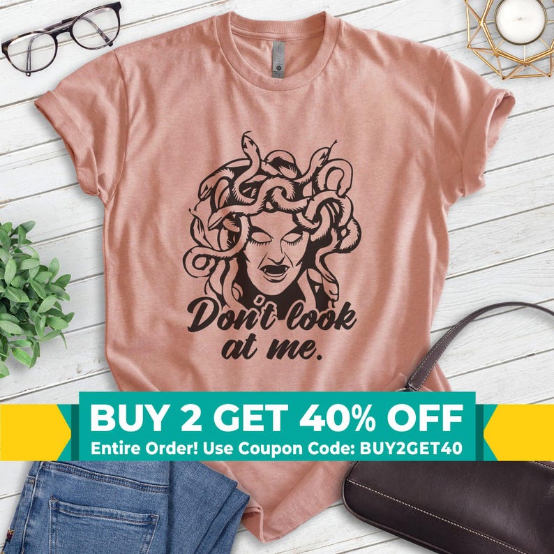 Pu&ograve; includere: T-shirt rosa chiaro con grafica nera di Medusa e la scritta "Don't look at me." La maglietta ha girocollo e maniche corte.