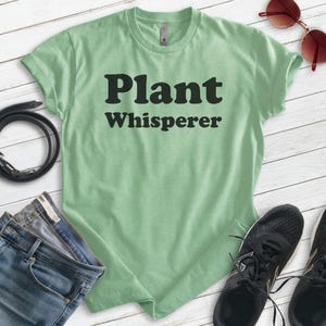 May include: A light green t-shirt with the text "Plant Whisperer" in black letters. The shirt is made of a soft, comfortable material and has a relaxed fit.