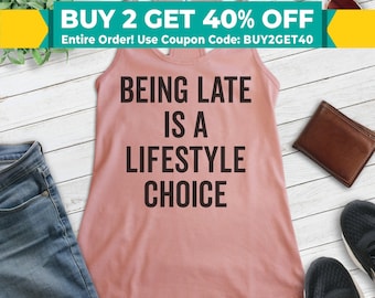 May include: A dusty rose racerback tank top with the text "BEING LATE IS A LIFESTYLE CHOICE" in black. The image includes a pair of sunglasses, a wallet, a pair of sneakers, and a pair of jeans.