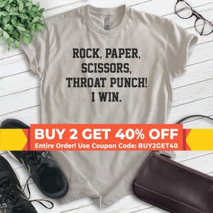 May include: A light gray t-shirt with black text that reads "ROCK, PAPER, SCISSORS, THROAT PUNCH! I WIN."  The t-shirt is laying on a wooden surface with a pair of black shoes and a brown purse. A banner at the bottom of the image reads "BUY 2 GET 40% OFF Entire Order! Use Coupon Code: BUY2GET40".