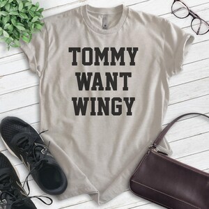 May include: A light beige t-shirt with the text "TOMMY WANT WINGY" in bold, black letters. The shirt is displayed on a white wooden surface, with black sneakers, a brown purse, and eyeglasses in the background.