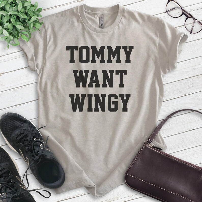 May include: A light beige t-shirt with the text "TOMMY WANT WINGY" in bold, black letters. The shirt is displayed on a white wooden surface, with black sneakers, a brown purse, and eyeglasses in the background.