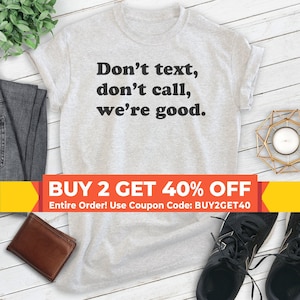 May include: A gray t-shirt with the text "Don't text, don't call, we're good." printed in black. The shirt is laying flat on a wooden surface with a pair of black sneakers, a brown leather wallet, and a gold candle holder.