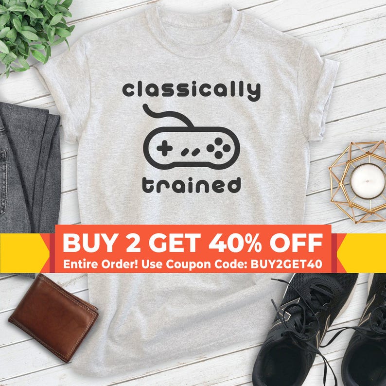 May include: A gray t-shirt with the text "classically trained" and a black and white illustration of a video game controller. The text "BUY 2 GET 40% OFF" is on a yellow and red banner below the shirt. The text "Entire Order! Use Coupon Code: BUY2GET40" is below the banner.