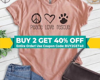 May include: A peach-colored t-shirt with a peace sign, heart, and paw print graphic. The text "Peace Love Rescue" is printed below the graphic. The t-shirt is displayed on a wooden surface with a pair of blue jeans and a brown purse.